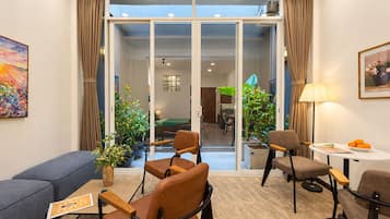 Una sala moderna con un sofá, dos sillones, una mesa de centro y una mesa de comedor con una lámpara. Hay plantas en macetas y una vista al área de comedor a través de puertas de vidrio.