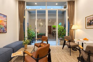 Un salon moderne comprenant un canapé, deux fauteuils, une table basse et une table à manger avec une lampe. On y trouve également des plantes en pot et, grâce à des portes vitrées, on peut apercevoir l’espace repas.