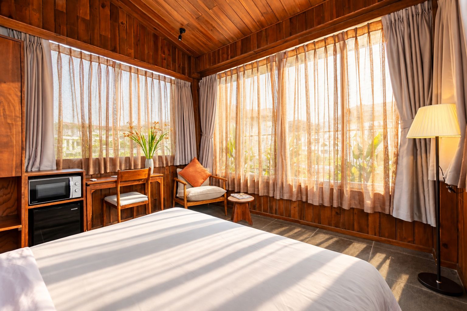 A room with a bed, a desk with a chair, a lamp, and a view of greenery outside.