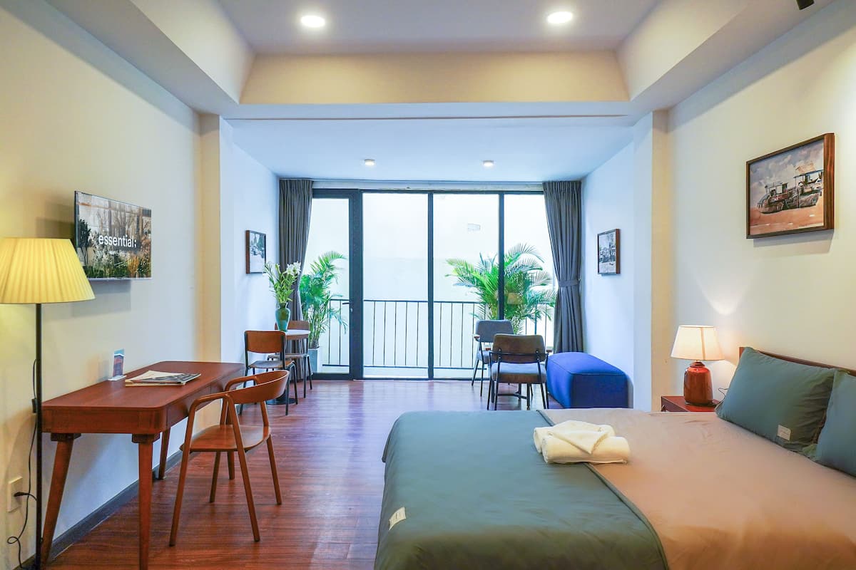 Habitación de hotel con cama, escritorio con sillas, balcón con plantas y un cuadro en la pared.