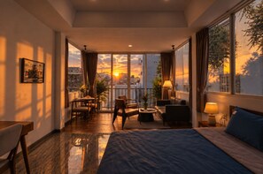 A hotel room with a large bed, a seating area with a table and chairs, a balcony with a view of the sunset, and a painting on the wall. - Nexus House Cay Diep - Near Independence Palace (Ho Chi Minh City)