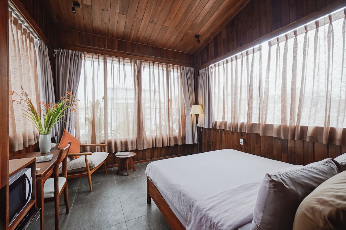 Una habitación con techo de madera, una cama con ropa blanca, un escritorio con silla y una mesita con lámpara.