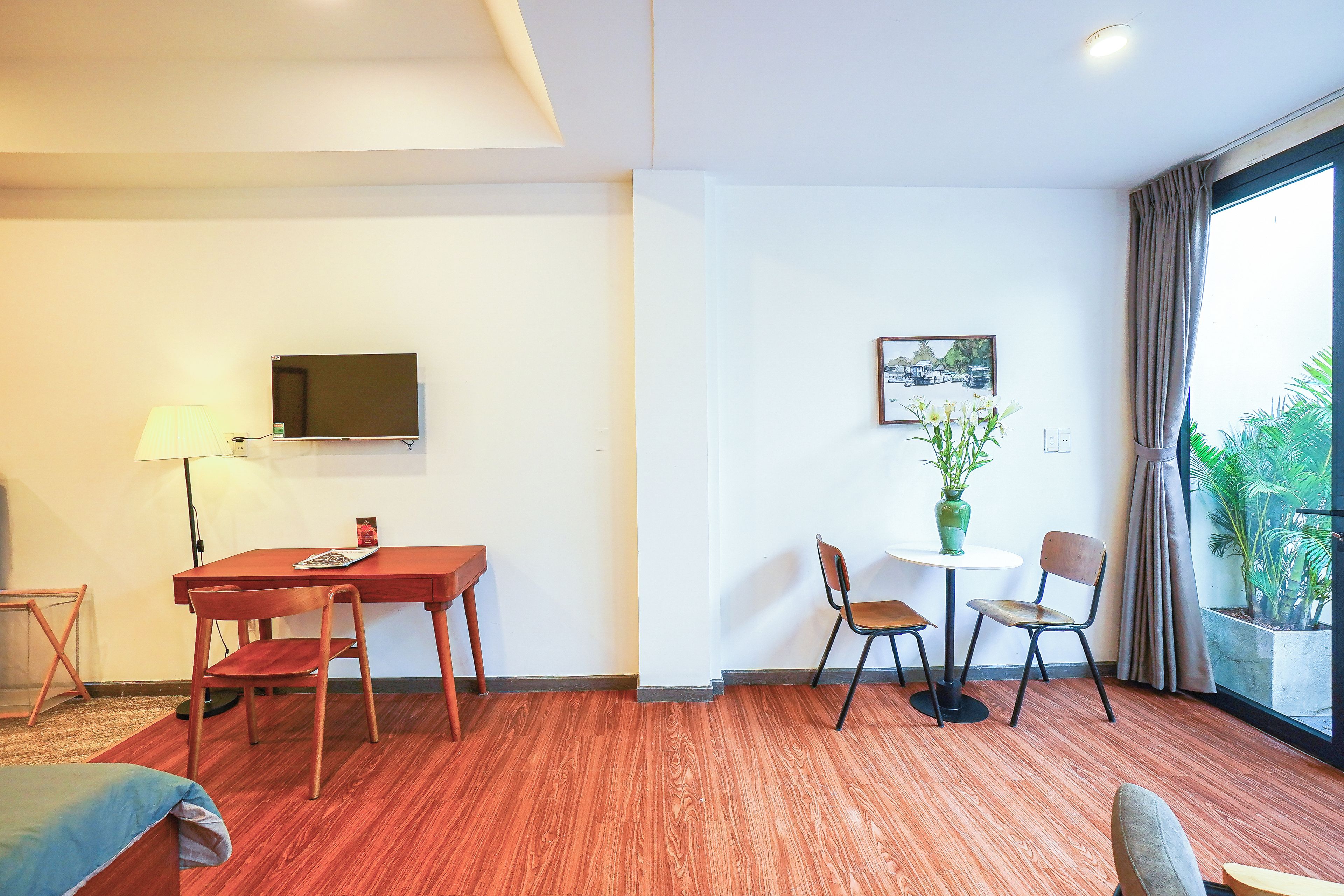 A hotel room with a wooden floor, a TV mounted on the wall, a dining table with chairs, a small table with a vase of flowers, and a window with curtains.