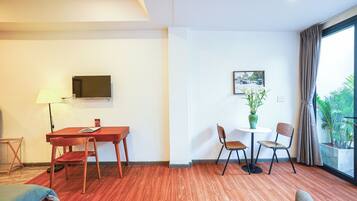 A hotel room with a wooden floor, a TV mounted on the wall, a dining table with chairs, a small table with a vase of flowers, and a window with curtains.
