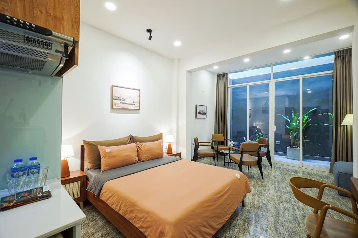 Habitación de hotel con cama, mesita de noche, lámpara y vistas al exterior a través de amplias ventanas.
