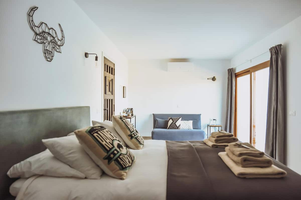 A hotel room with a large bed, a sofa, a wall-mounted air conditioner, and a window with curtains.