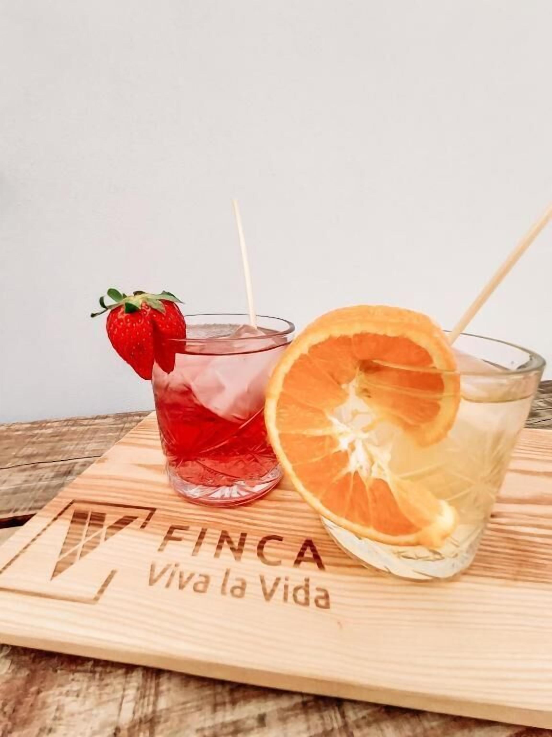 Two glasses of red beverage with a strawberry and an orange slice, on a wooden board with "FINCA Viva la Vida" engraved.