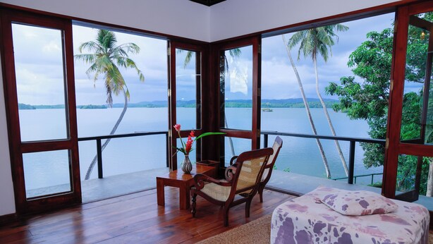 Quarto com piso de madeira, cama com colcha estampada, cadeira, mesinha com um vaso de flores e janelas amplas com vista para um corpo de água e palmeiras.