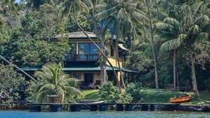 Uma casa de dois andares com uma varanda, rodeada por palmeiras e um pequeno barco na água.