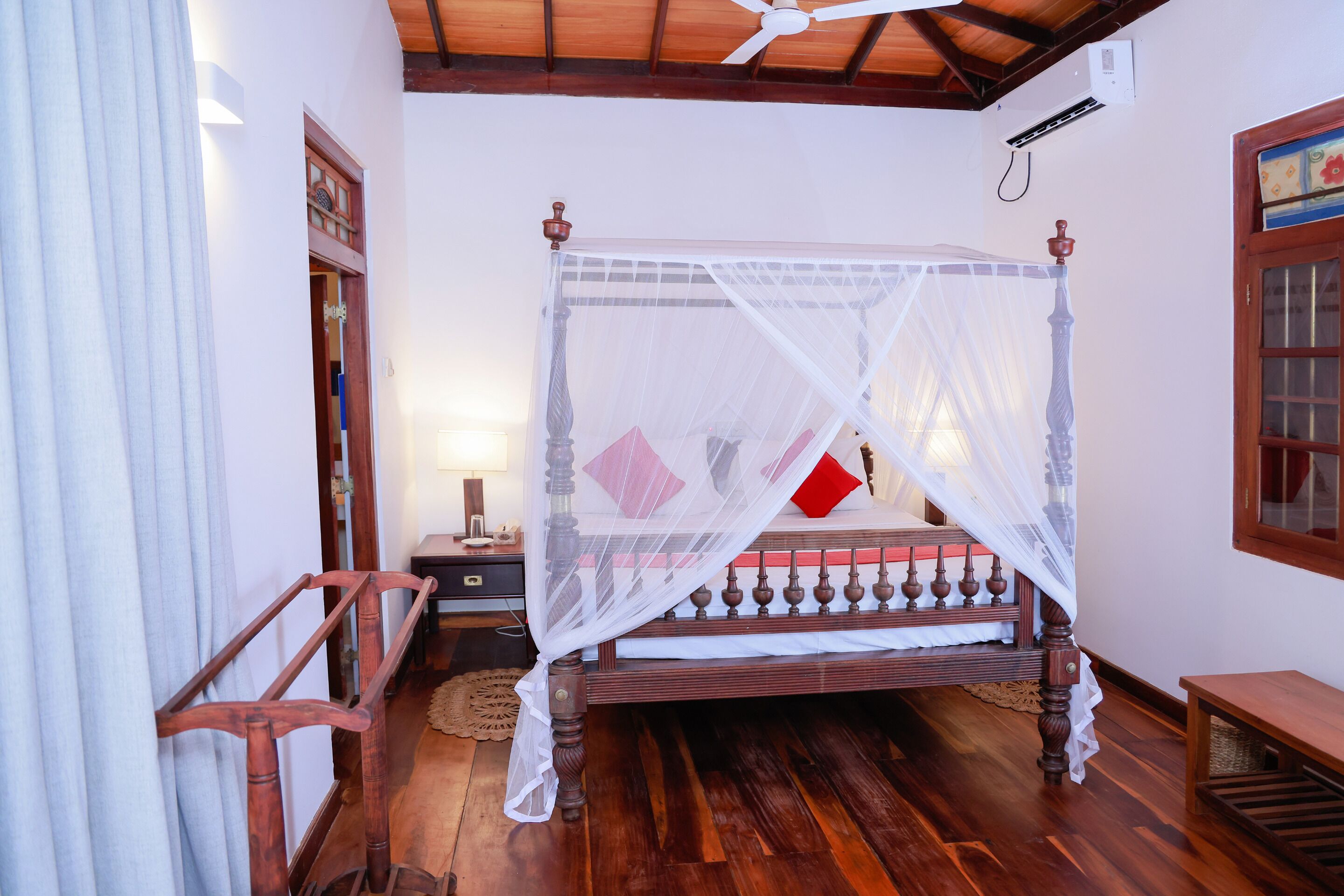 Una cama con dosel sobre cuatro columnas, dosel blanco, suelo de madera, ventilador de techo y mesita de noche con lámpara.