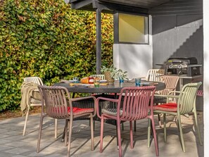 A round outdoor dining table with chairs, a grill, and a hedge in the background.