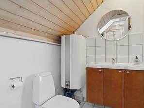 Uma casa de banho moderna com sanita branca, teto de madeira, lavatório branco com espelho e parede revestida a azulejos brancos.
