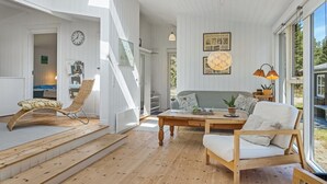 Un vaste espace de vie doté d’un plancher en bois, d’un canapé, d’une table à manger et d’un fauteuil en osier. Une grande fenêtre offre une vue sur l’extérieur.
