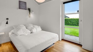 Une chambre moderne avec un grand lit, un luminaire au plafond, un panneau décoratif fixé au mur et une vue sur un jardin verdoyant, visible à travers une porte-fenêtre coulissante.