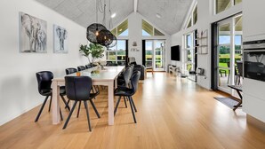 Un vaste espace dédié à la salle à manger, avec une longue table et des chaises, une cuisine moderne dotée d’un grand four et de grandes fenêtres offrant une vue sur l’extérieur.