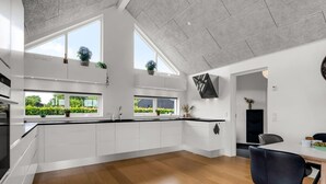Une cuisine moderne avec des armoires blanches, un comptoir noir et un plancher en bois. On y trouve des fenêtres avec des plantes sur le rebord, ainsi qu’un coin repas avec une chaise noire.