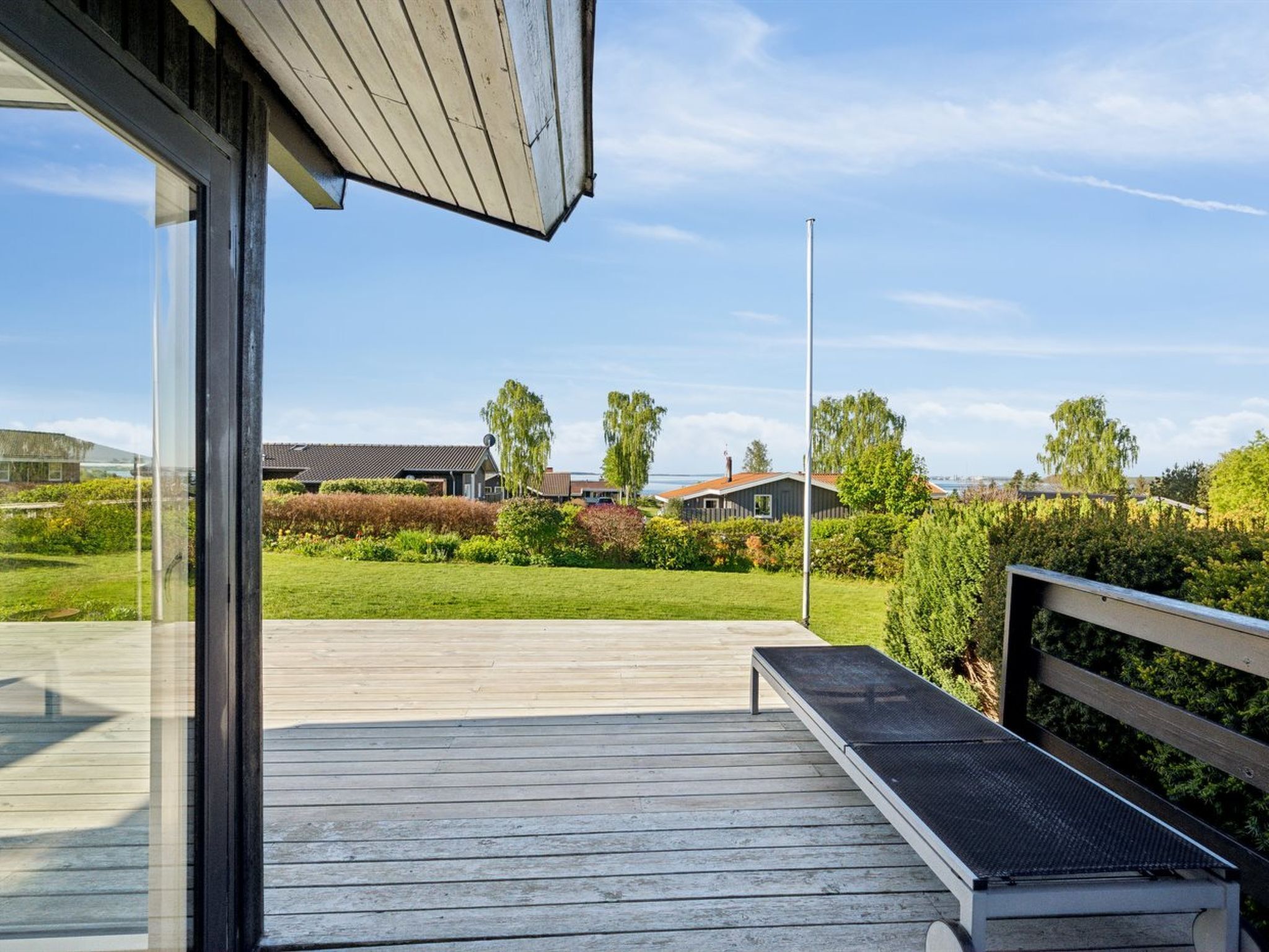 Een houten terras met een bank, een glazen deur en uitzicht op een groen landschap met huizen en bomen.
