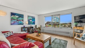 A modern living room with a red sofa, a wooden coffee table, and a bookshelf. There is a large window with a view of houses and greenery. - "Hilppa" - 400m from the sea by Interhome (Harboøre)