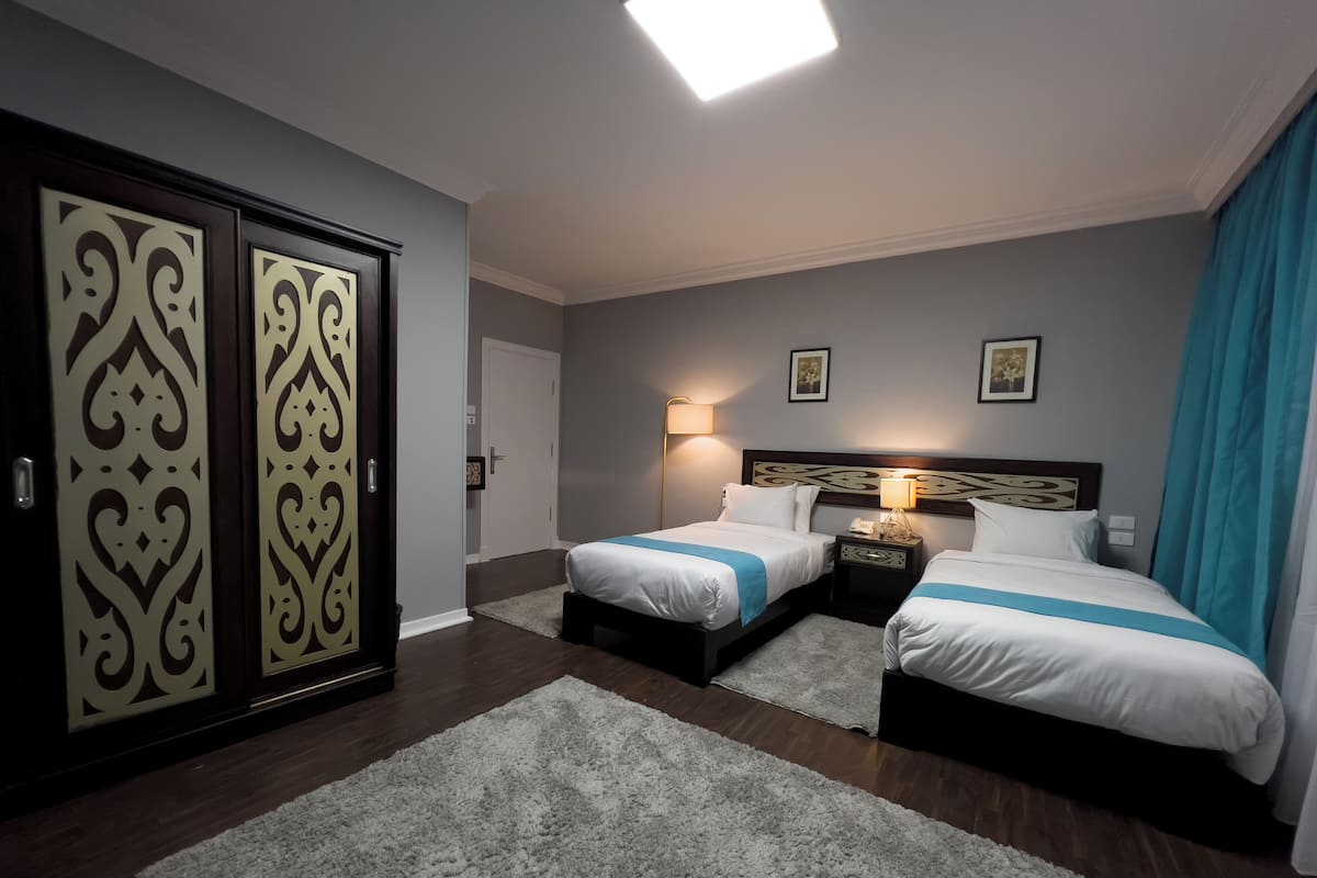 A hotel room with two beds, a nightstand, a lamp, and a large window with curtains.