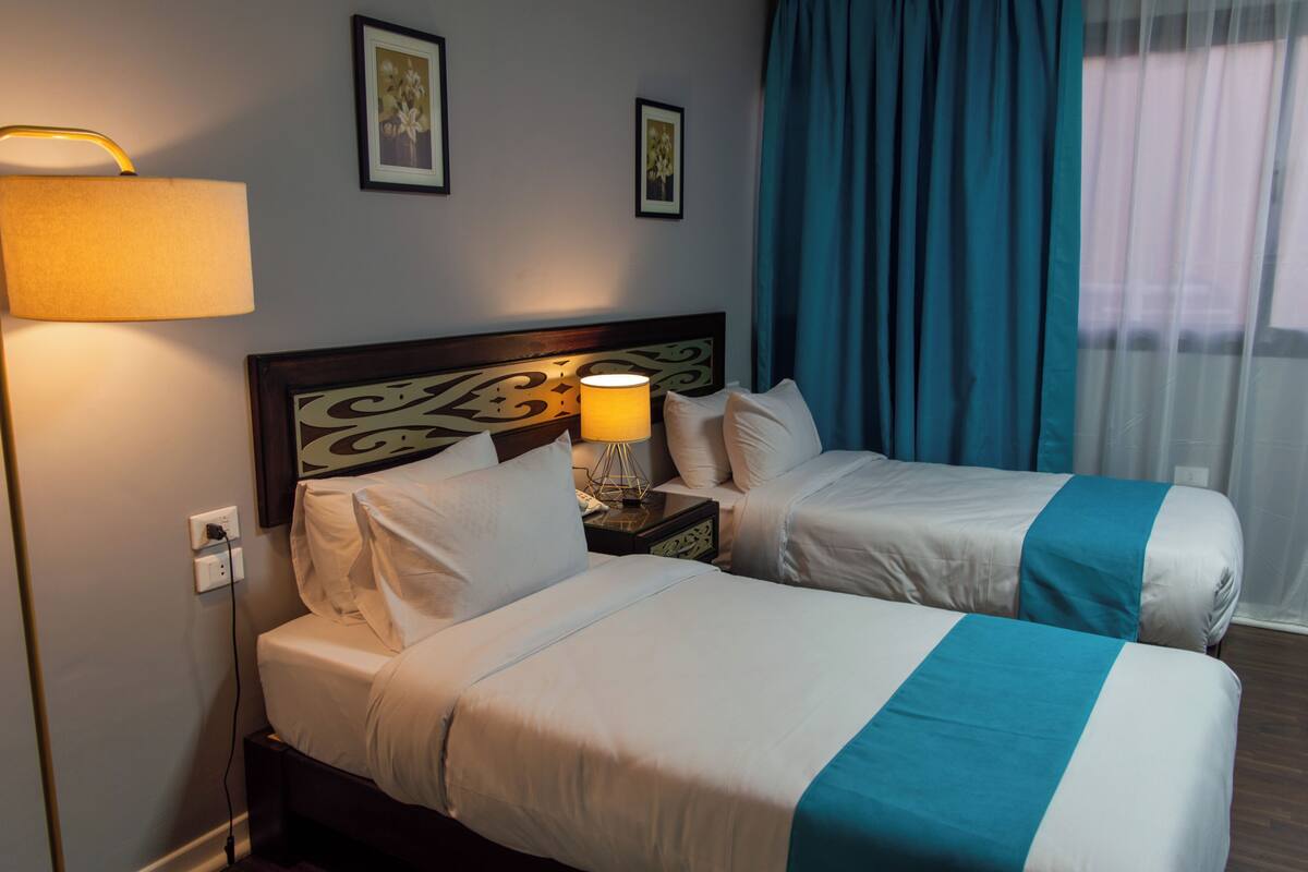 A hotel room with two beds, a nightstand with a lamp, and a window with curtains.
