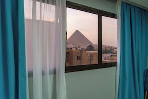 A room with a window view of a pyramid, curtains, and a glimpse of the surrounding buildings.