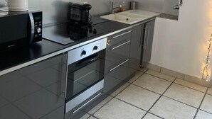 A compact kitchen with a microwave, coffee maker, and oven. There is a small sink and a shelf with decorative items. - Charming house (Remoulins)