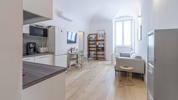 A modern kitchen with a microwave, toaster, and coffee maker. There is a dining area with a table and chairs, and a sofa. A bookshelf and a window with natural light are also visible.