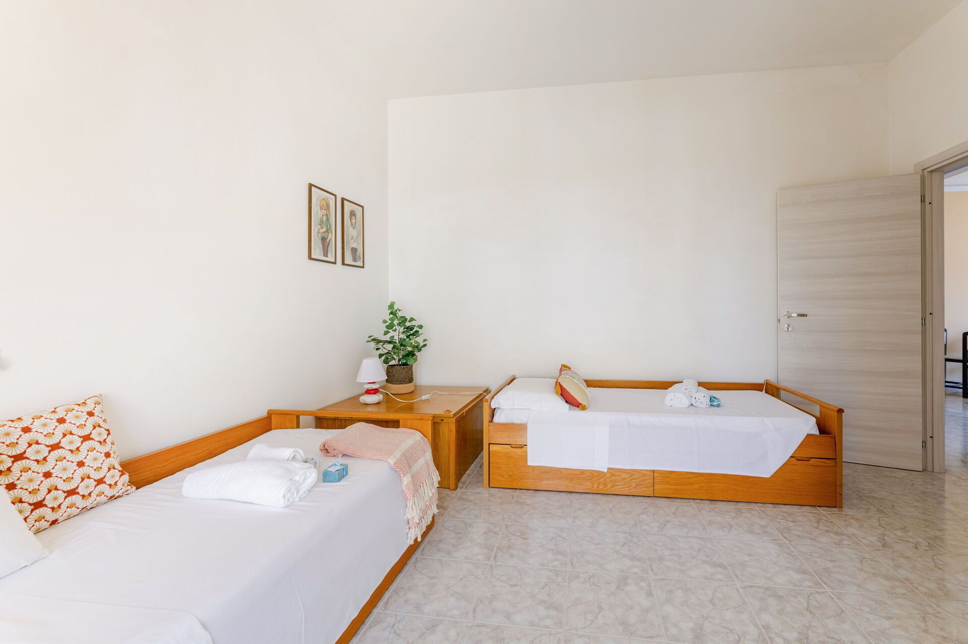 Une chambre d’hôtel avec deux lits simples, une table de chevet avec une lampe, une petite plante et une porte sur le côté droit.