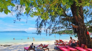 Uma praia com águas azul-claras, pessoas nadando e tomando sol, e uma grande árvore oferecendo sombra.