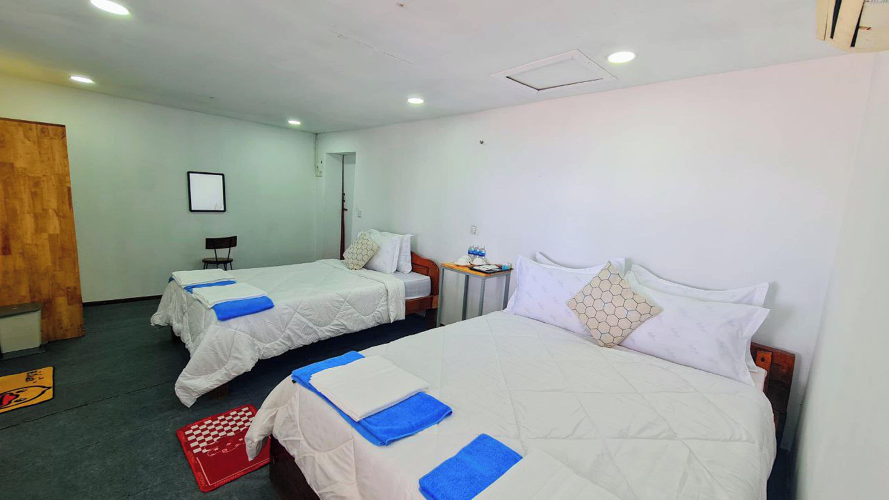Two single beds with white bedding and blue towels, a small table with a water bottle, and a red checkered mat on the floor.