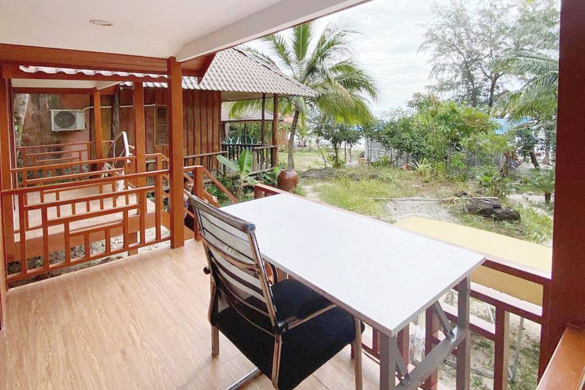 A room with a wooden floor, a table, and a chair. There is a view of palm trees and greenery outside.