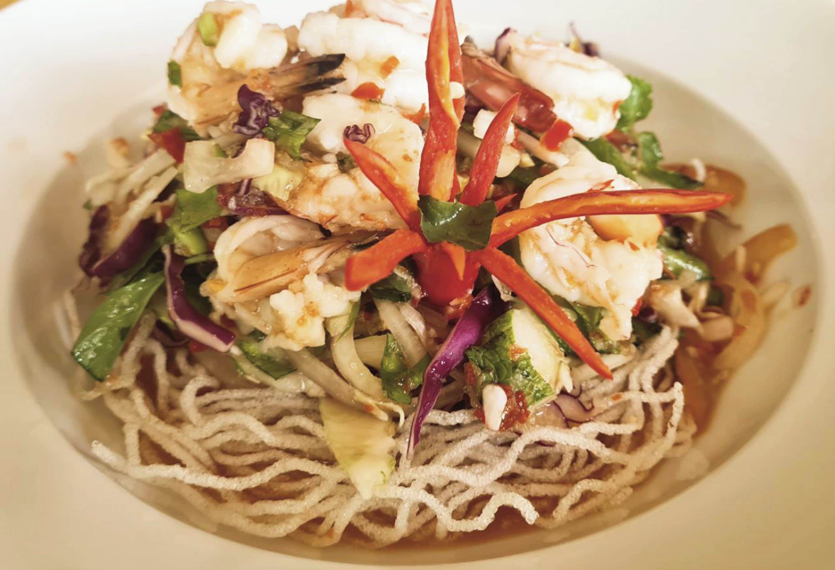 Un plato con fideos, camarones, verduras y un tallo de chile decorativo.