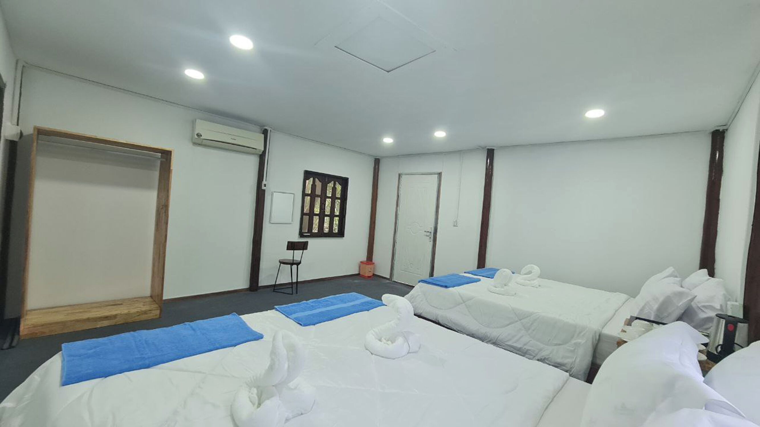 Habitación de hotel con dos camas, una puerta de madera, un equipo de aire acondicionado, una mesita con una silla y una ventana con marco de madera.