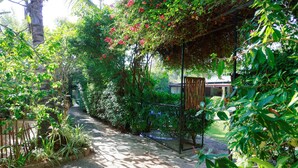 Un vialetto nel giardino, con una pergola ricoperta di piante rampicanti fiorite, immerso nel verde e con un piccolo edificio sullo sfondo.