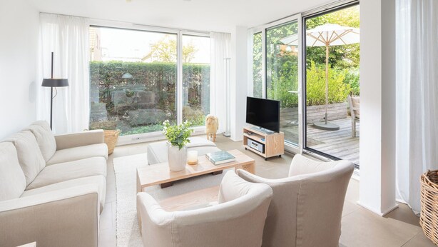 A modern living room with a sofa set, a coffee table with decorative items, a flat-screen TV on a wooden stand, and large windows offering a view of a garden and outdoor seating area. - // Pudelsand - Eins // (Norderney)