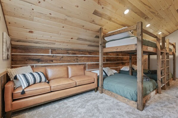 Una cama litera con estructura de madera y ropa de cama verde, un sofá de cuero marrón con almohadones a rayas, y una pared blanca con detalles de paneles de madera.