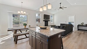Una cocina moderna con encimera de granito, armarios oscuros y una isla central. Hay un comedor con una mesa de madera y bancos, un sofá oscuro y un ventilador de techo.