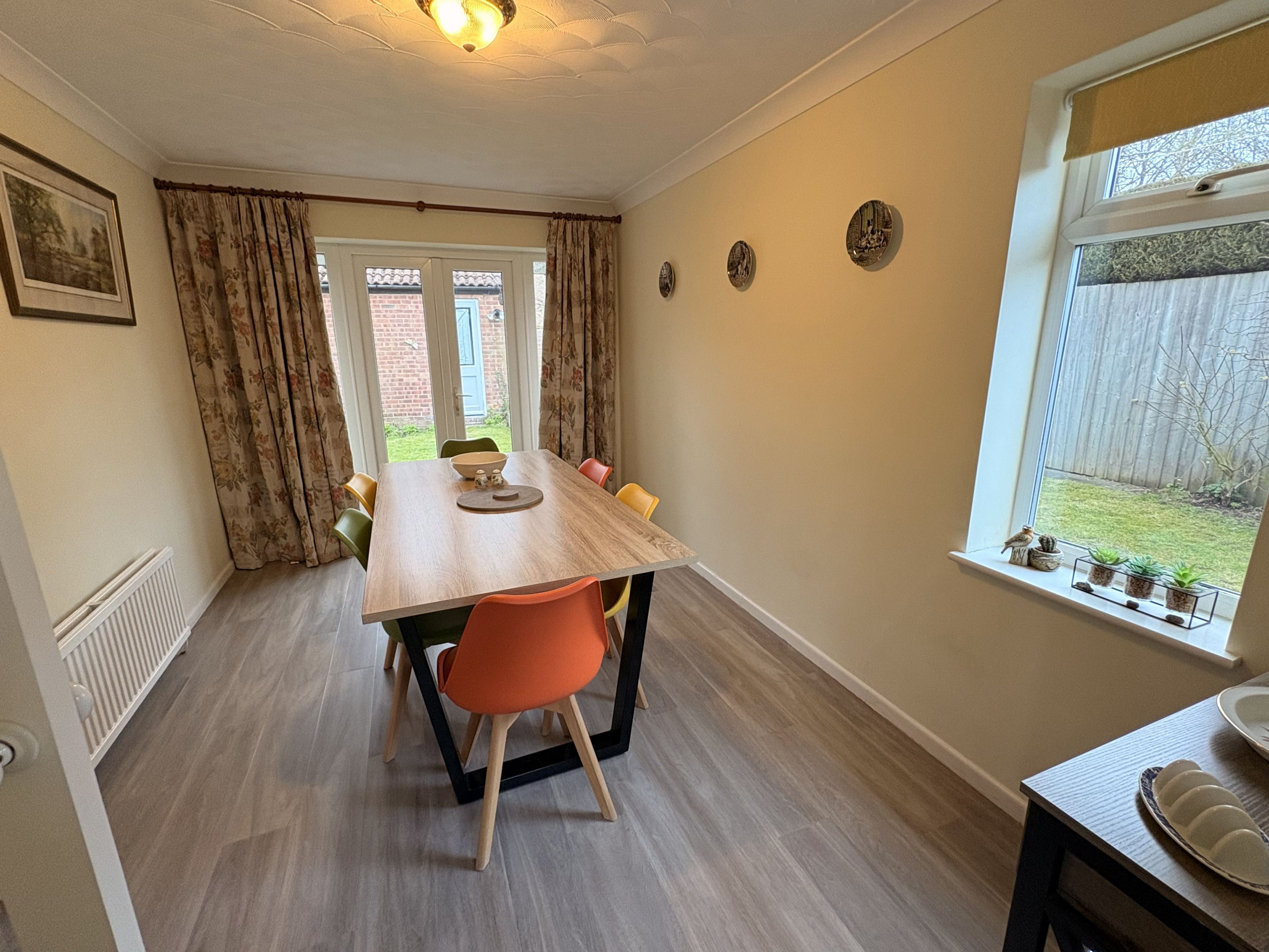 A dining area with a wooden table, orange chairs, a radiator, and a window with a view of a garden.