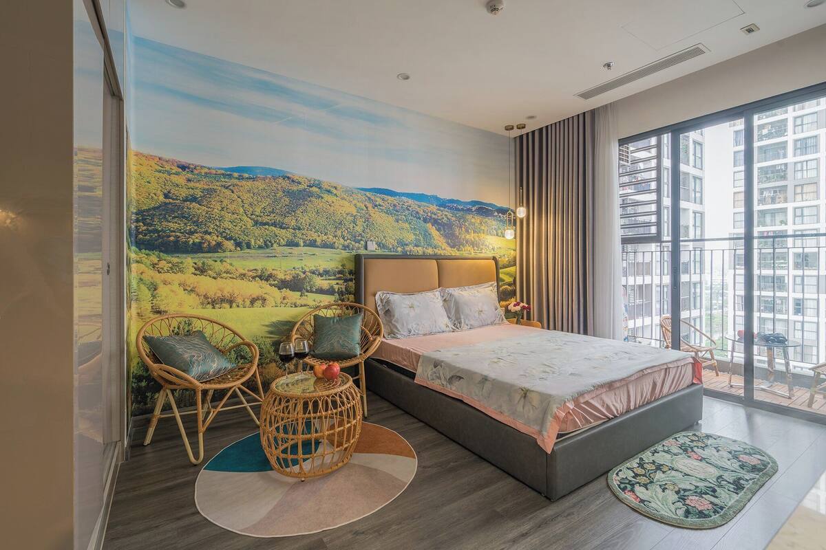Quarto com uma cama grande, duas cadeiras de vime, uma mesa redonda pequena e vista para a cidade através de amplas janelas.
