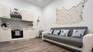 A modern kitchen with white cabinets, a built-in oven, and a small island. There is a grey sofa with patterned cushions and a decorative wall hanging.