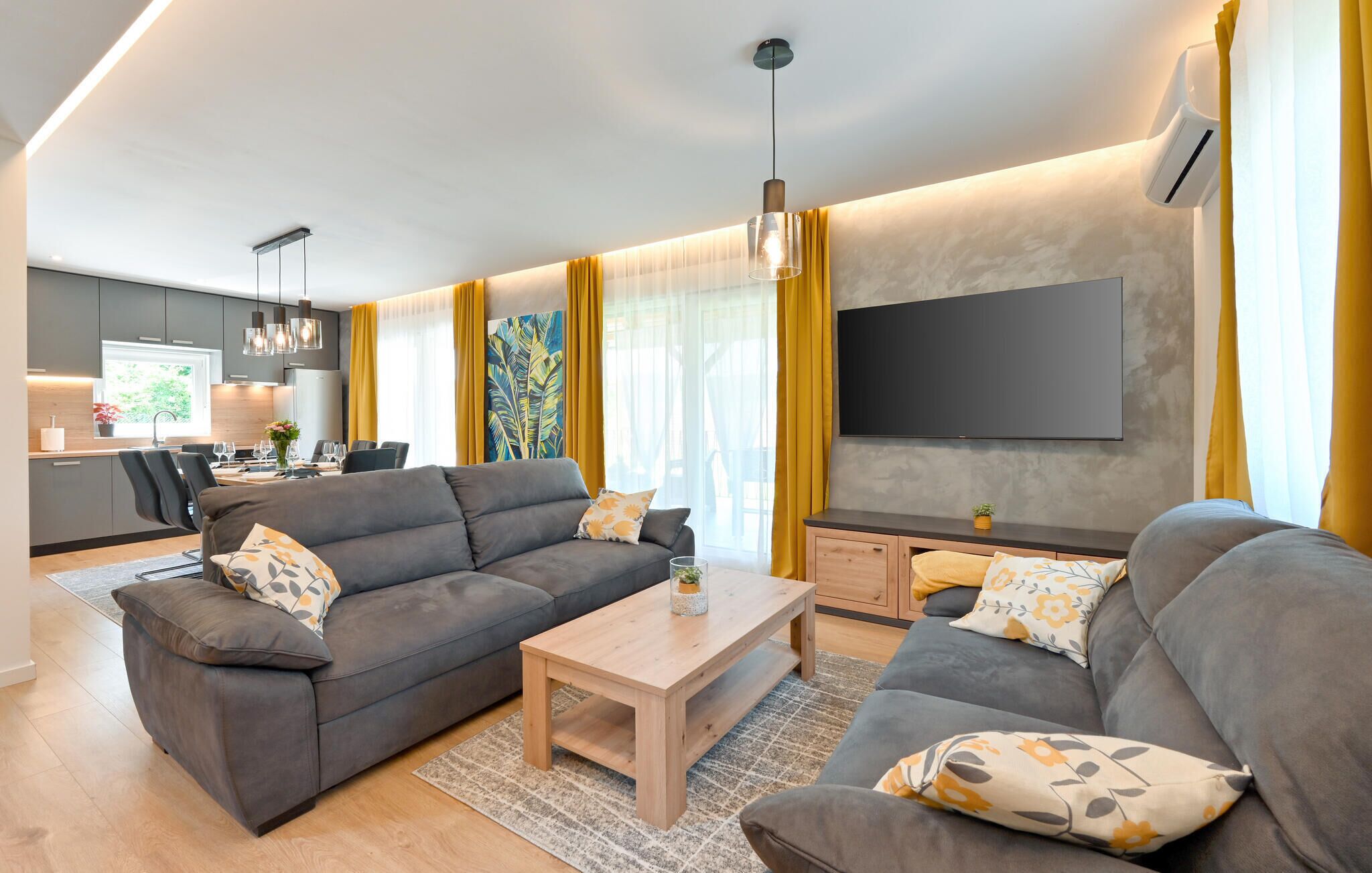 A modern living room with grey sofas, a wooden coffee table, and a flat-screen TV mounted on the wall.