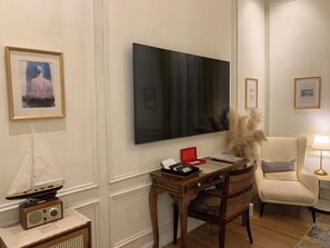 A room with a large flat-screen TV mounted on the wall, a wooden desk with a red box, a chair with a checkered cushion, and a small ship radio on a side table. - The Atlantic Langkawi (Langkawi)