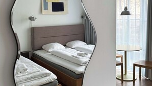 Une chambre moderne dotée d’un miroir au design original, d’un lit soigneusement fait, d’une petite table de chevet et offrant une vue sur l’extérieur grâce à de grandes fenêtres.