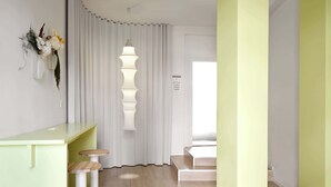 Une chambre d’hôtel moderne avec un bureau et des tabourets de couleur vert clair, un rideau blanc et un panneau indiquant les services disponibles, tels que la blanchisserie et le service de bagagerie.