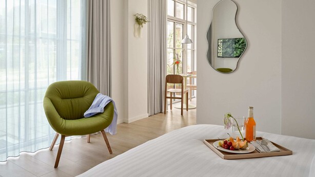 Habitación de hotel moderna con una cama grande, un sillón verde, una zona de comedor con mesa y sillas, y un espejo con una planta.