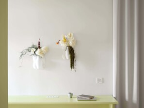 A wall with two white vases containing flowers and greenery, a small table with a book and a cup, and a light switch on the wall. - Studio Apartment for Solo Travelers (København)