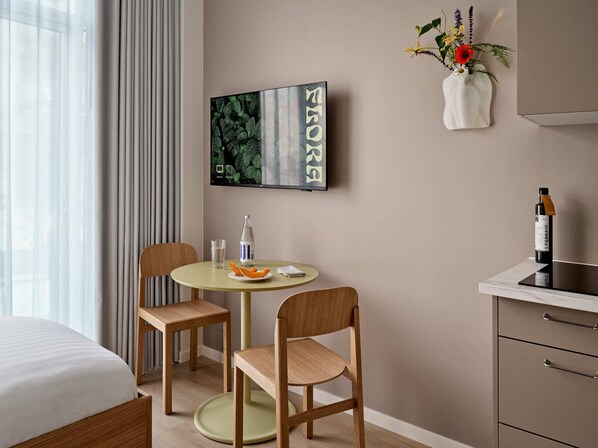 A modern hotel room with a bed, a round table with chairs, a small table with a bottle and a glass, a painting on the wall, and a vase with flowers.