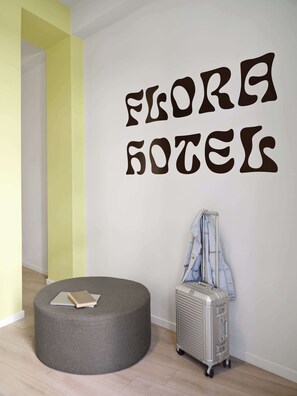 Ein Zimmer mit grauem Hocker, einem Koffer und einer Jacke, die auf einem Gestell hängt. An der Wand steht in stilvoller Schrift "FLORA HOTEL".