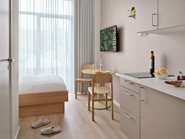 A modern hotel room with a bed, a desk with a laptop, a chair, a small table with a bowl of fruit, and slippers on the floor.