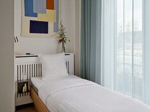 A neatly made bed with a white comforter, a white pillow, and a small bedside table with a glass of water and a bottle. There is a decorative wall hanging above the bed and a window with light blue curtains.